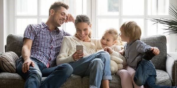 Happy family of four sitting on a sofa with a woman holding a mobile, capturing a moment of joy.